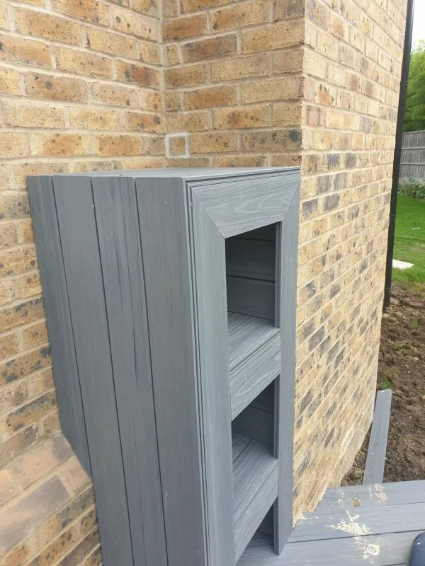 Grey decking boards with built-in storage shelves beside a brick wall